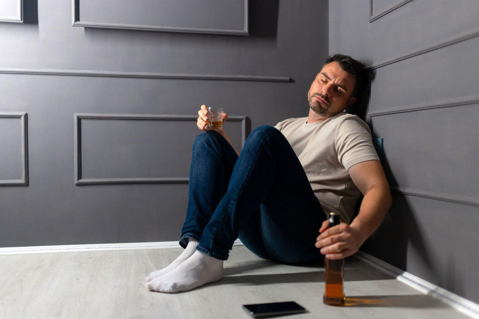 A man sitting slumped against a wall on the floor holding a glass and a bottle of liquor.