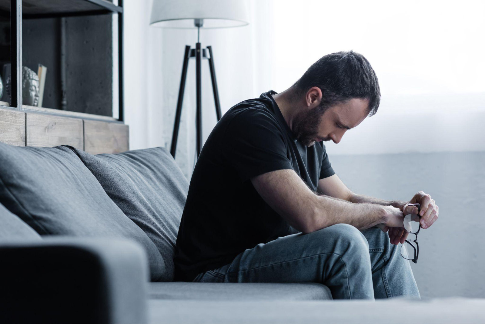 man-struggling-with-addiction-sitting-alone A middle-aged man sitting alone on a gray sofa in a living room, leaning forward in distress while holding his glasses.