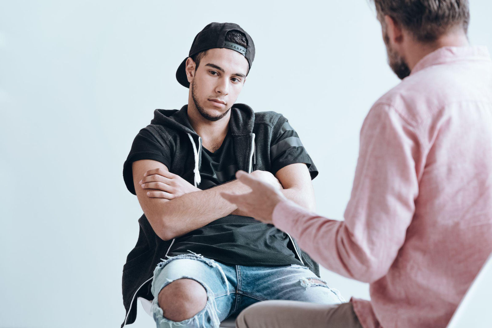A young man sitting with arms crossed and a hesitant expression while someone speaks to him.