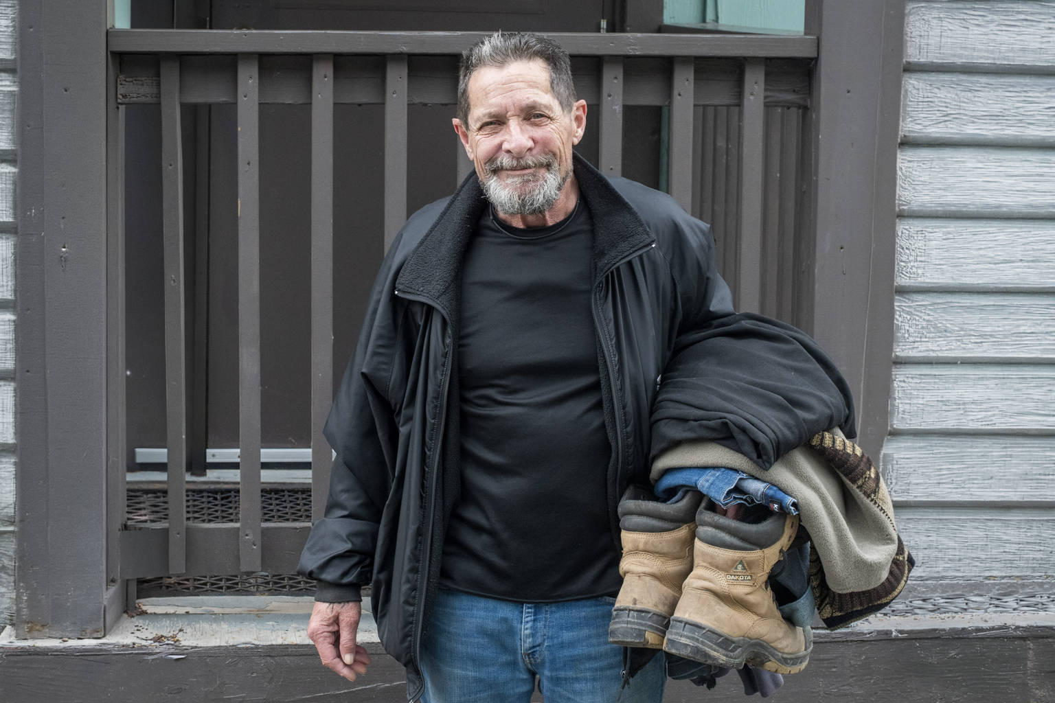 Older smiling man standing outside holding his folded clothes and work boots, representing a fresh start in addiction recovery.
